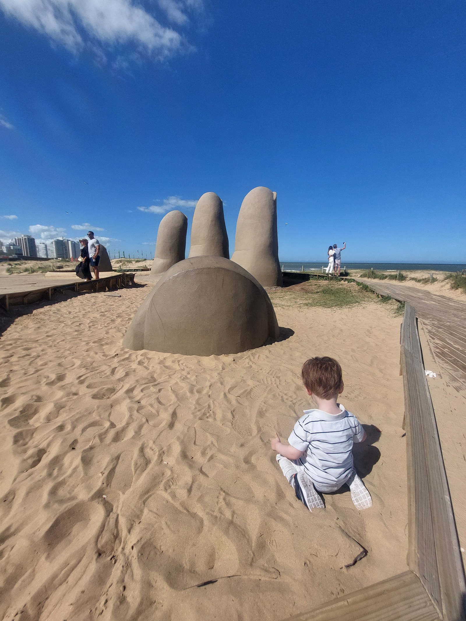 Lolo sitzt im Sand vor der Mano-Skulptur in Punta del Este (von hinten)